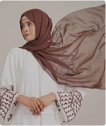 Woman wearing a brown hijab and white outfit with patterned sleeves against a neutral background